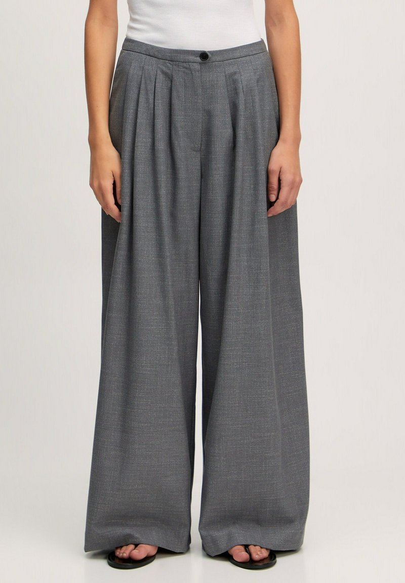 Pantalons larges gris avec des plis, fabriqués à partir d'un tissu texturé. Dotés d'une taille avec fermeture à bouton. Pas de poches visibles.