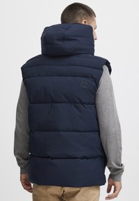 Navy blue puffer vest with a high hood, featuring a quilted pattern and logo detail on the back, worn over a grey long-sleeve shirt.
