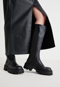 Svarte knelhøye støvler med en tykk såle, glatt skinntekstur, glidelås på siden og minimalistisk design, kombinert med et svart skjørt med splitt.