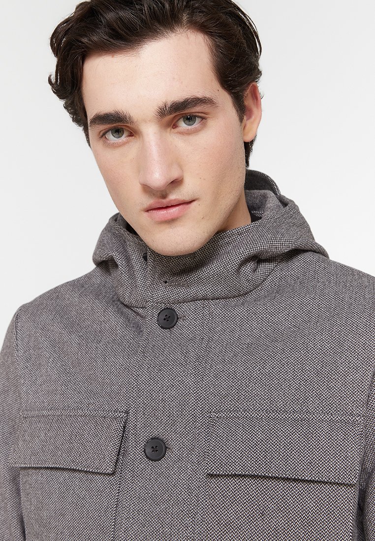 Jonge man met donker haar in een grijze hakket-jacket met een capuchon en twee knoopzakken op de borst tegen een witte achtergrond.