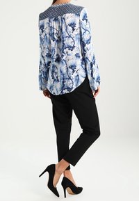 Blouse bleue à motifs floraux avec un empiècement texturé bleu marine, associée à un pantalon noir ajusté et des chaussures à talons hauts noires.
