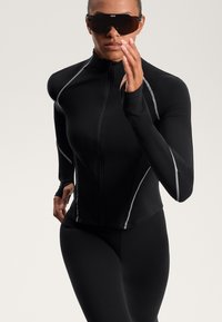 Fekete sportdzseki cipzárral az elején, hosszú ujjakkal és fényvisszaverő szürke díszítéssel, fitted fekete leggings-szel és napszemüveggel párosítva.