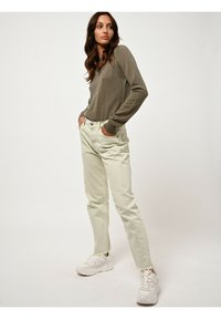 Olivengrüner Pullover mit V-Ausschnitt, kombiniert mit hellbeigen Jeans. Modell trägt weiße Sneakers. Glatte Texturen und taillierte Passform.