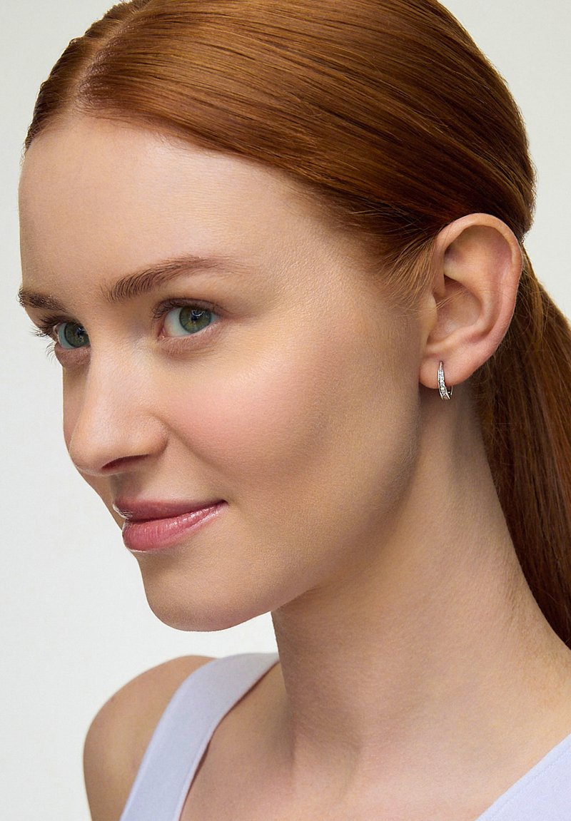 Srebrne kolczyki w kształcie kółek o teksturowanej powierzchni i subtelnym blasku, noszone przez modelkę z długimi rudymi włosami i wyraźnymi zielonymi oczami.