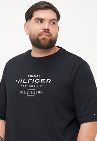 Czarny t-shirt z bawełny z białą grafiką logo, która na przodzie brzmi "TOMMY HILFIGER NEW YORK CITY Est. 1985". Okrągły dekolt, krótkie rękawy.