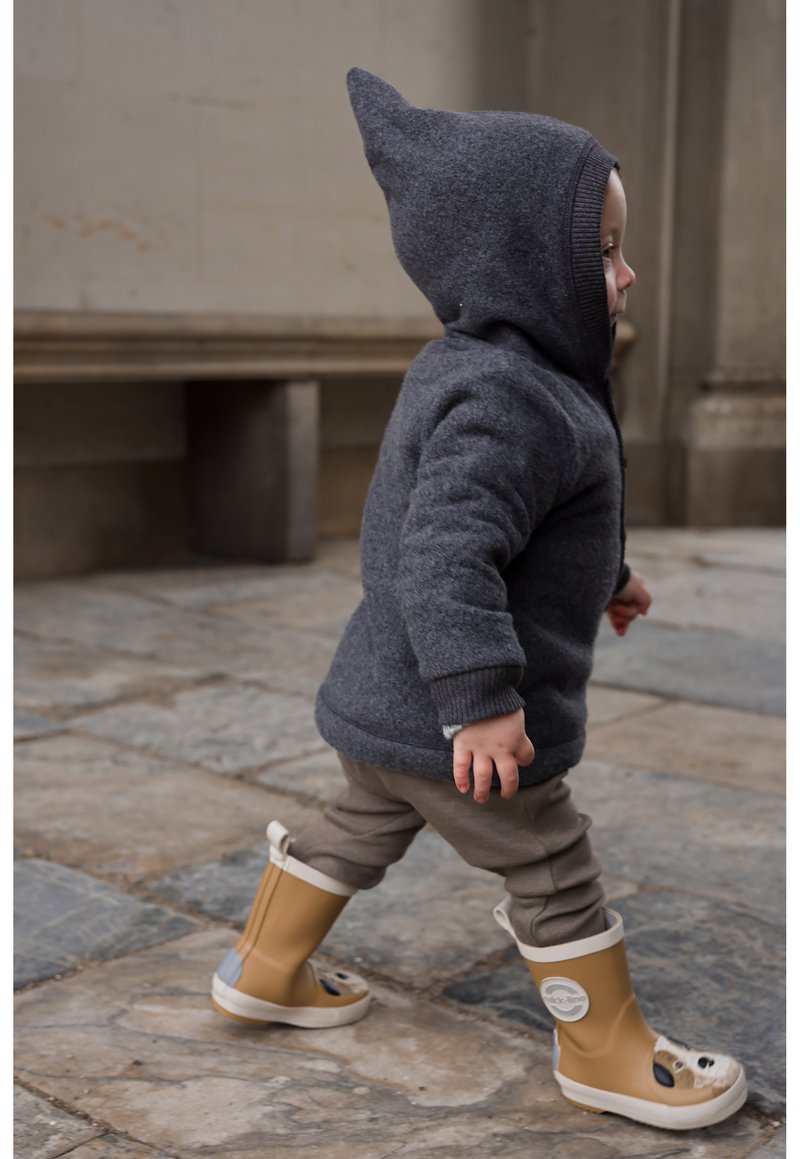 Sweat à capuche en polaire grise avec capuche pointue, bottes imperméables couleur tan avec accents blancs, pantalon beige, marchant sur un pavé en pierre.