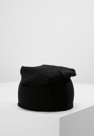 Zwarte wollen baret met een zachte, platte bovenkant, rustend op een wit voetstuk tegen een lichtgrijze achtergrond.