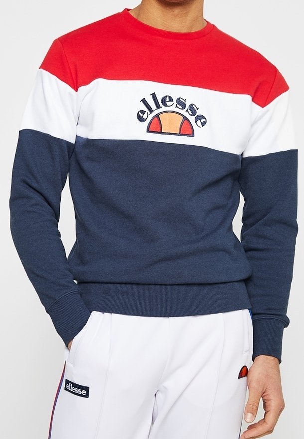 Homme portant un sweat-shirt color-block rouge, blanc et marine avec le logo « ellesse », associé à un pantalon de sport blanc avec des écussons logo.