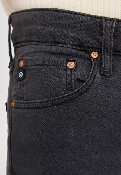 Jeans din denim negru cu un design fitted, cu accesorii din cupru și o etichetă mică de marcă pe buzunar. Detaliu texturat pe material vizibil.