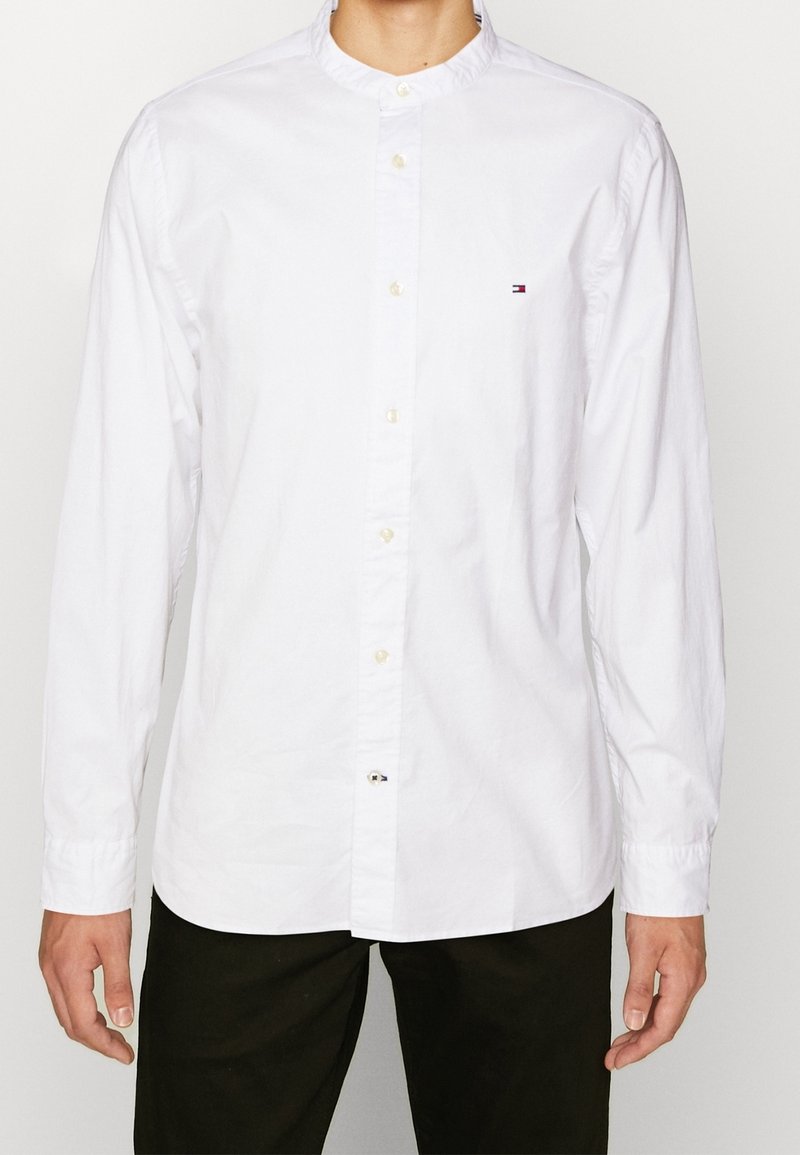 Homme portant une chemise blanche à manches longues avec un petit logo sur la poitrine et un pantalon noir.