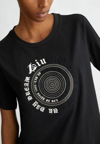 Maglietta nera con design grafico circolare, testo bianco e pattern a pois. Maniche corte e scollatura rotonda. Tessuto di cotone morbido.