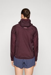 Bordeauxroter leichter Jacke mit Kapuze, aus dehnbarem Material, leicht strukturiertem Finish und Logo-Detail im Nackenbereich.