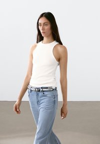 Hvid ærmeløs top med høj hals, parret med lysblå jeans og et dekorativt bælte. Glat tekstur og tætsiddende design.