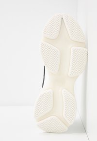 Semelle d'une basket claire avec des zones de grip texturées montrant un motif de bande de roulement en zigzag, positionnée verticalement sur un fond blanc.