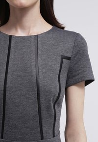 Grå klänning med kort ärm som har svarta accentlinjer fram och en sidficka, tillverkad av ett strukturerat tyg.