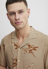 Camisa de botones de manga corta en color beige con patrones de hojas de palma marrones. Tejido ligero con un cuello relajado y una textura suave.