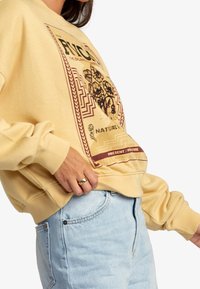 Sudadera amarilla de manga larga con un diseño gráfico impreso, cuello redondo y puños de canalé. El material parece suave y texturizado.