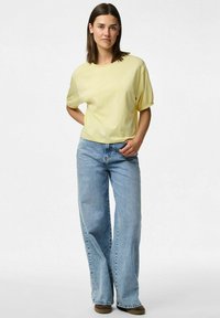 Gul t-shirt med kort ärm och lös passform, kombinerad med ljusblå vida jeans och bruna skor. Enkelt design utan ytterligare mönster.