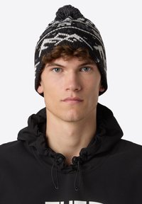 Berretto a maglia in bianco e nero con un motivo geometrico e un pompon in cima. Indossato con una felpa nera con colletto a cordino.