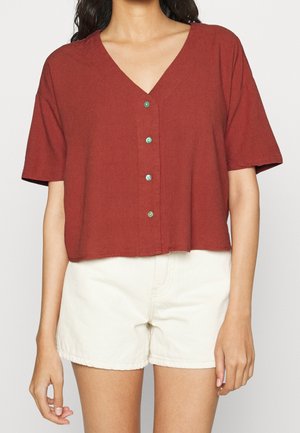 Frau trägt ein rostrotfarbenes, kurzärmeliges V-Ausschnitt-Button-up-Oberteil in Kombination mit hoch taillierten cremeweißen Shorts, dargestellt von den Schultern bis zur Mitte der Oberschenkel.