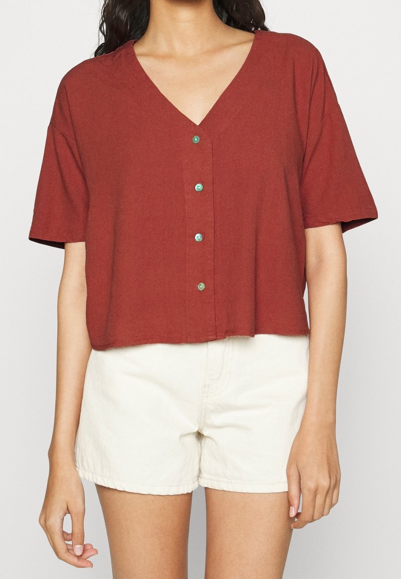 Femme portant un haut rouille à manches courtes avec boutons et col en V, associé à un short taille haute blanc cassé, visible des épaules jusqu'au milieu des cuisses.