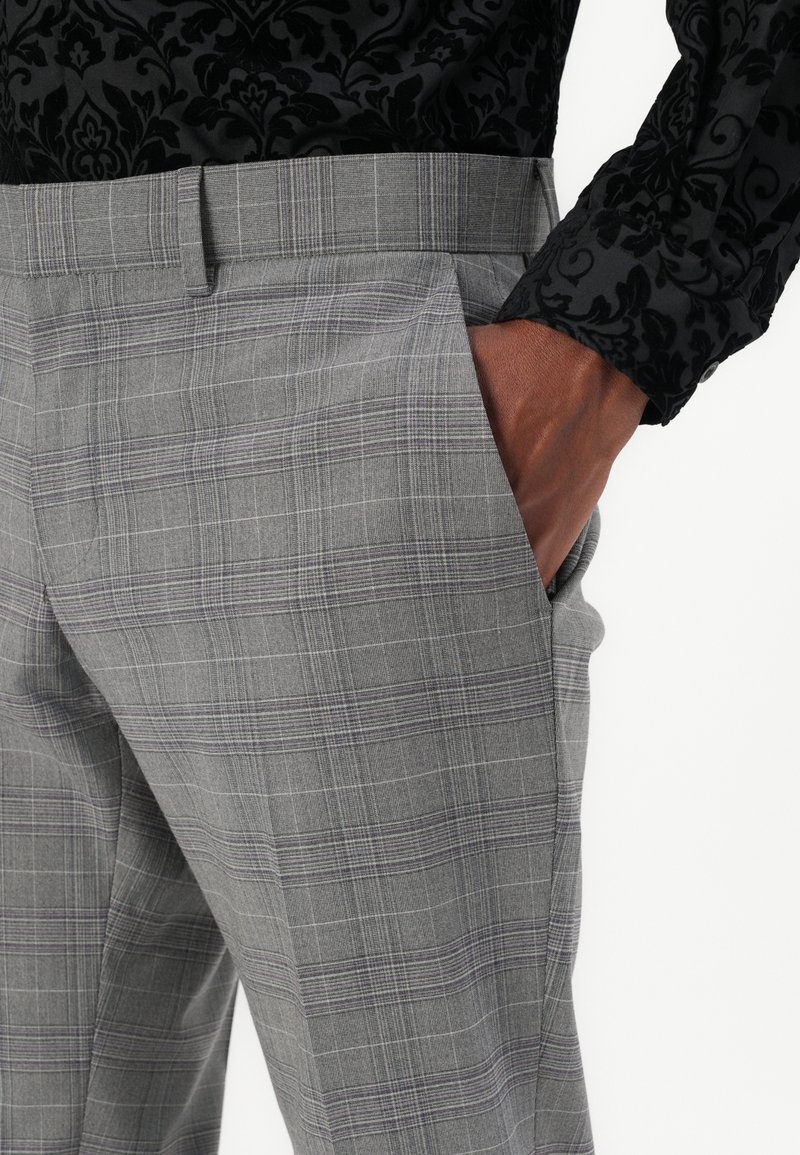 Grijze geruite dressbroek met een gladde textuur, voorzien van een subtiel ruitpatroon en zijzakken. Een gebogen hand rust in een zak.