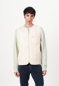 Chaleco acolchado beige con cuello redondo, cierre frontal de cremallera y un bolsillo en el pecho. Lleva puesto sobre una camisa de manga larga verde claro.