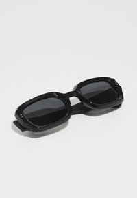Ochelari de soare negri cu un cadru gros, unghiular, lentile întunecate și accente metalice aurii pe partea din față. Fabricati din material plastic lucios.