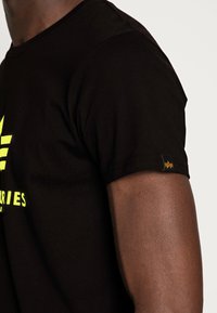 Black cotton t-shirt featuring yellow graphic print on the front, short sleeves, and a small branded tag on the left sleeve.