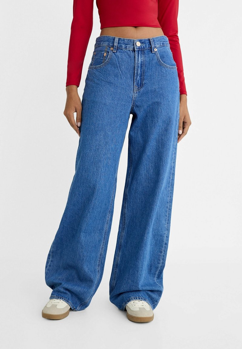 Personne portant un jean bleu ample taille haute, des baskets blanches et un top court rouge à manches longues, debout devant un fond uni.