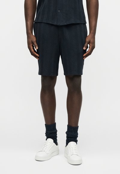 Personne portant un short texturé foncé, une chemise assortie, des chaussettes foncées froissées et des baskets blanches à lacets, debout devant un fond blanc uni.