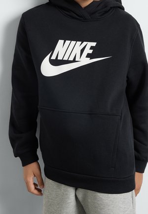 Osoba ubrana w czarną bluzę z kapturem Nike z dużym białym logo oraz szare spodnie dresowe, stojąca na tle jasnoszarego tła.