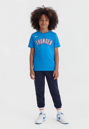 Enfant aux cheveux bouclés portant un t-shirt bleu Thunder, un pantalon de survêtement bleu marine, des chaussettes blanches avec des rayures orange, et des baskets blanches avec des accents bleus.