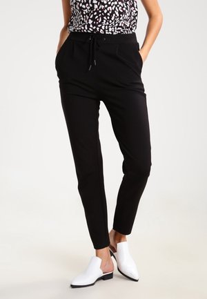 Kobieta ubrana w czarne spodnie zwężane z ściągaczem w talii, wzorzysty top bez rękawów oraz białe wsuwane buty na spiczastym nosku, stojąca z rękami w kieszeniach.