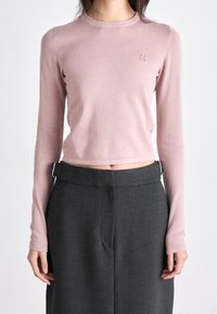 Kobieta nosząca dopasowany, jasnoróżowy crop top z długim rękawem oraz wysokie, ciemnoszare spodnie z bocznymi klamrami.