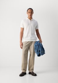 Witte geribbelde poloshirt, lichtbeige wijde broek en bruine enkellaarsjes. Blauwe denimjasje in de linkerhand gehouden. Effen achtergrond.