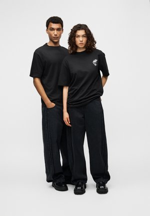 Tricouri negre supradimensionate cu un mic desen floral alb, asortate cu pantaloni negri din denim cu picioare largi și pantofi negri masivi.