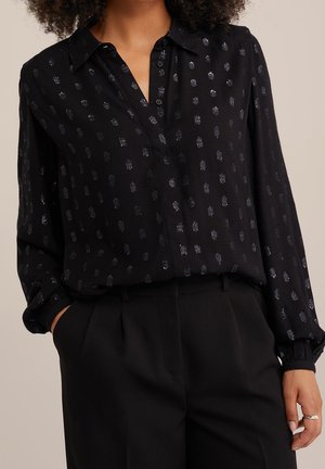 Blouse noire à manches longues avec un délicat motif de petits points argentés brillants, boutonnée sur le devant, portée rentrée dans un pantalon noir taille haute.