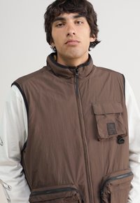 Brown vest made of lightweight fabric, featuring a high collar, zippered front, and two front pockets with zip closures and flap details.