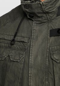 Olivgrön jacka i texturerat tyg med hög krage, knapprader, och två bröstfickor, med svarta detaljer och dragsko.