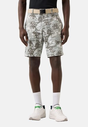 Férfi, szürke és fehér pálmaleveles terepmintás rövidnadrágban, bézs övvel, fekete pólóban, fehér zokniban és fehér sportcipőben zöld fűzővel, egyenesen áll.