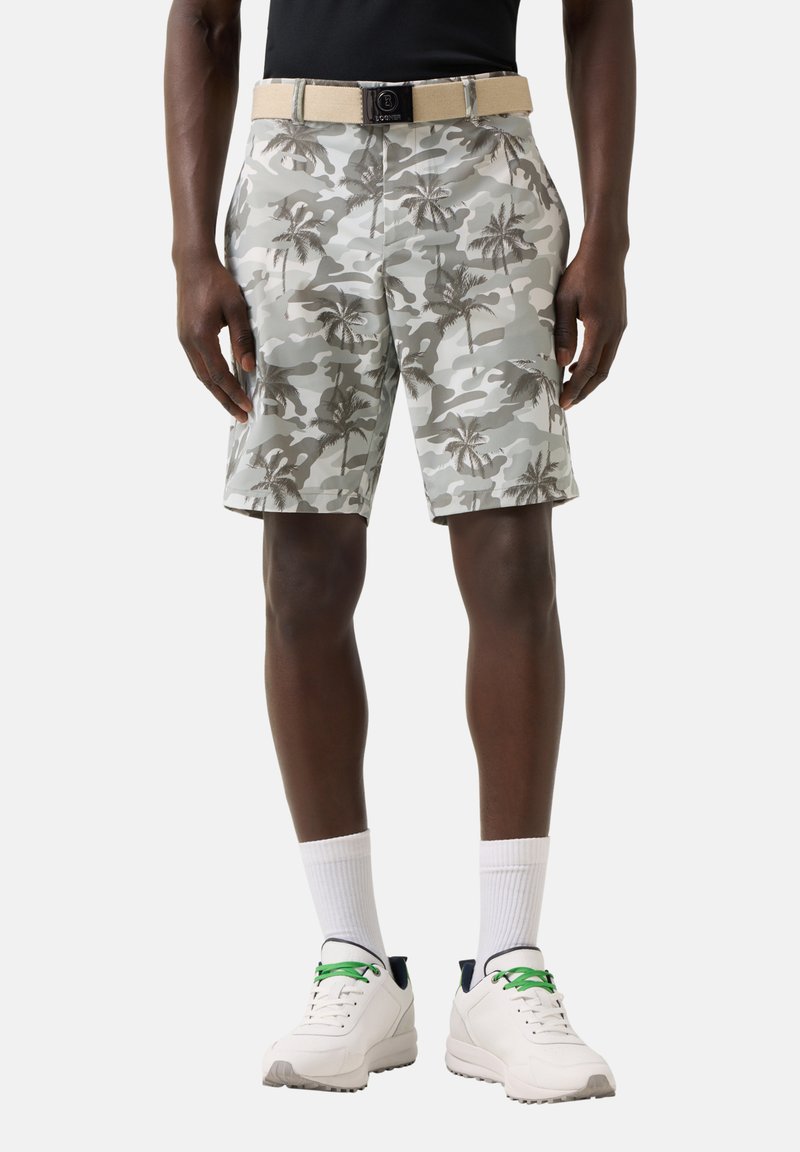 Άνδρας που φοράει γκρι και λευκά σορτς με καμουφλάζ φοίνικα, μπεζ ζώνη, μαύρο πουκάμισο, λευκές κάλτσες και λευκά αθλητικά παπούτσια με πράσινα κορδόνια, στέκεται ευθεία.