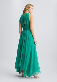 Zelena chiffon haljina s visokim izrezom, otvorenim leđima i lepršavom suknjom. Ima vezan struk i slojevitu teksturu. Elegantna i lagana dizajn.