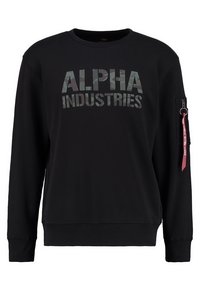 Sudadera negra con "ALPHA INDUSTRIES" en estampado de camuflaje en el pecho y una etiqueta roja "REMOVE BEFORE FLIGHT" en el bolsillo con cremallera de la manga izquierda.