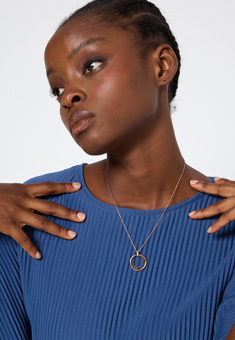 Collier en argent avec un pendentif circulaire arborant un texte gravé, porté sur un haut à rayures bleues. Le pendentif se détache sur le tissu.