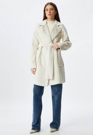 White coat with a soft, textured collar, long sleeves, and a tied belt. Features two front pockets and is paired with blue denim jeans.