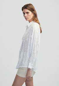 Chemise blanche, légère, à manches longues avec un design texturé et effiloché et des poignets à boutons ; portée par-dessus une mini-jupe de couleur claire.