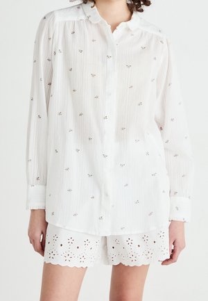 White long-sleeve blouse with small floral patterns paired with white scalloped eyelet shorts on a standing person.