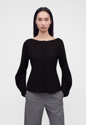 Jeune femme aux longs cheveux foncés portant un haut noir boutonné avec des manches larges et un pantalon gris à carreaux, debout devant un fond uni.
