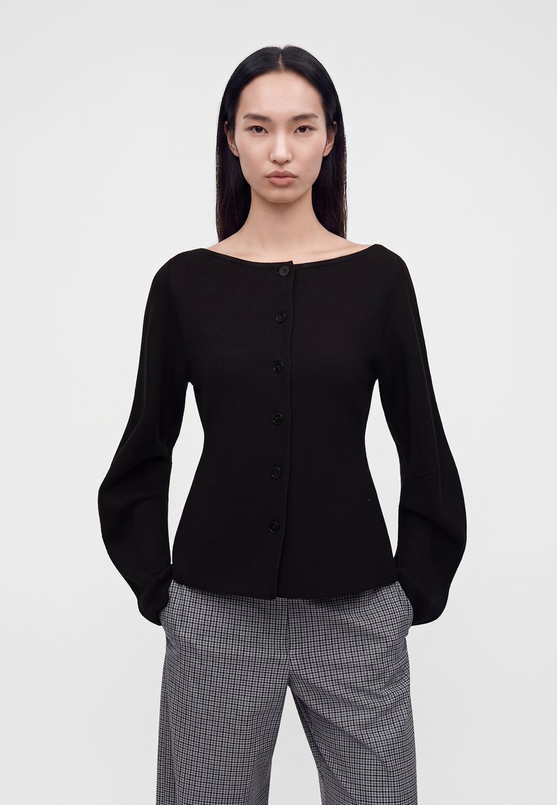 Jeune femme aux longs cheveux foncés portant un haut noir boutonné avec des manches larges et un pantalon gris à carreaux, debout devant un fond uni.